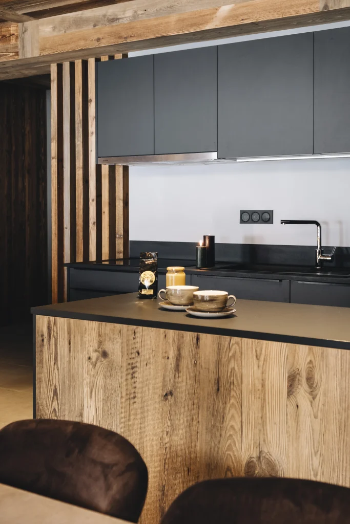 Vue de l'îlot central en bois massif texturé (style vieux bois brossé) et plan de travail anthracite mat d'une cuisine sur-mesure contemporaine Maison Morel. Ambiance chalet moderne chaleureux.