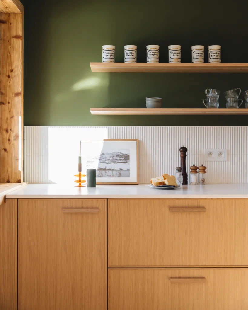 Vue détaillée d'une cuisine sur-mesure contemporaine avec meubles bas en chêne texturé plan de travail blanc mat et crédence texturée. Ambiance Japandi et naturelle par Morel. Sur le plan : pain, photo encadrée et décoration. Étagères en bois assorties.