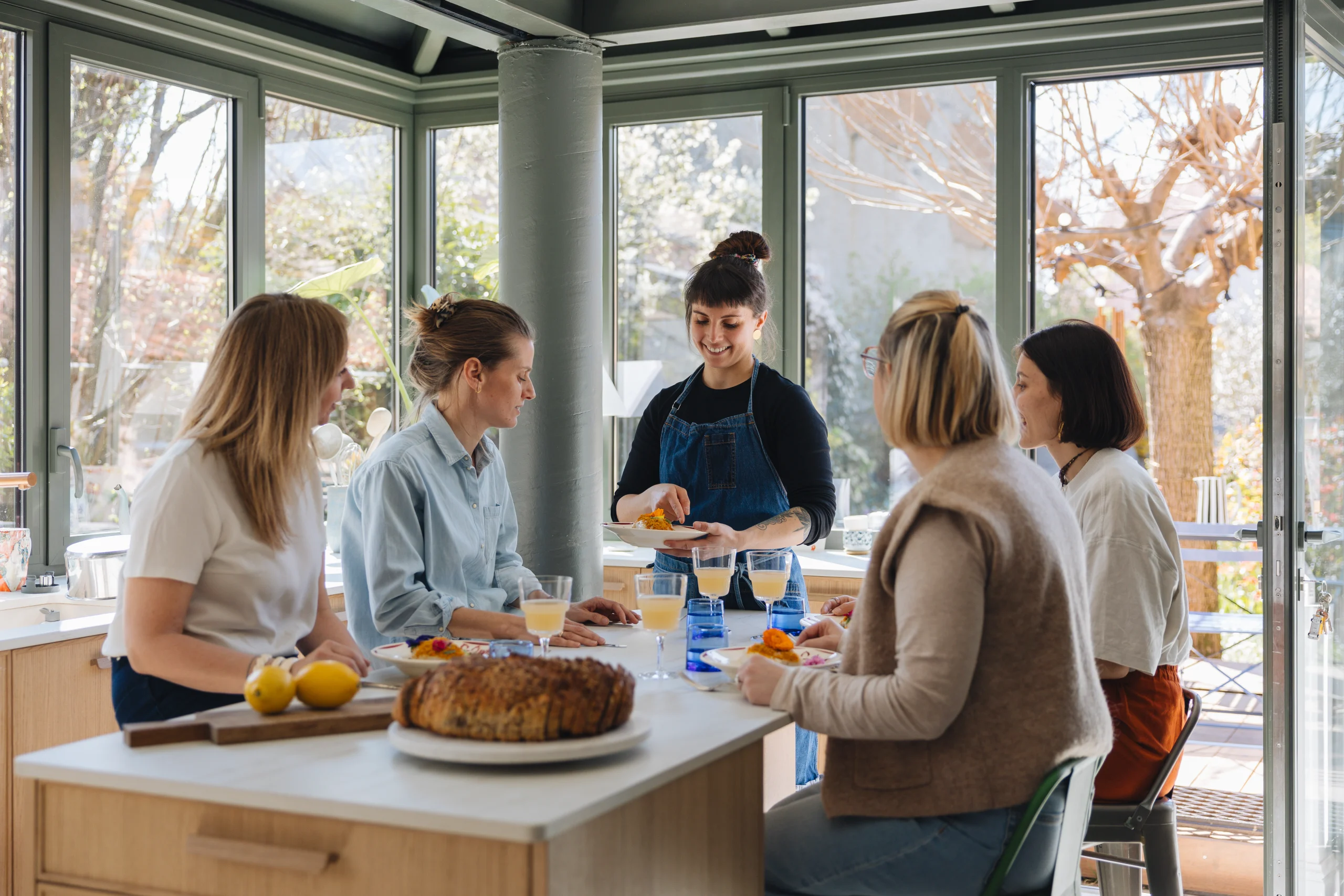 Aménagement Morel d'une cuisine lumineuse avec îlot en bois clair et plan de travail blanc, où des femmes partagent un repas.