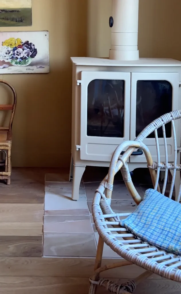 Coin salon avec poêle en fonte crème posé sur une dalle en céramique, entre parquet bois et fauteuil en rotin