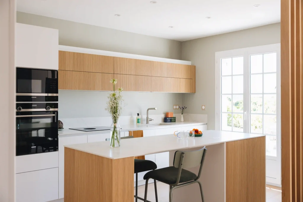 Cuisine bois moderne Morel mêlant façades blanches mates et bois clair strié, avec îlot-bar et électroménager noir encastré