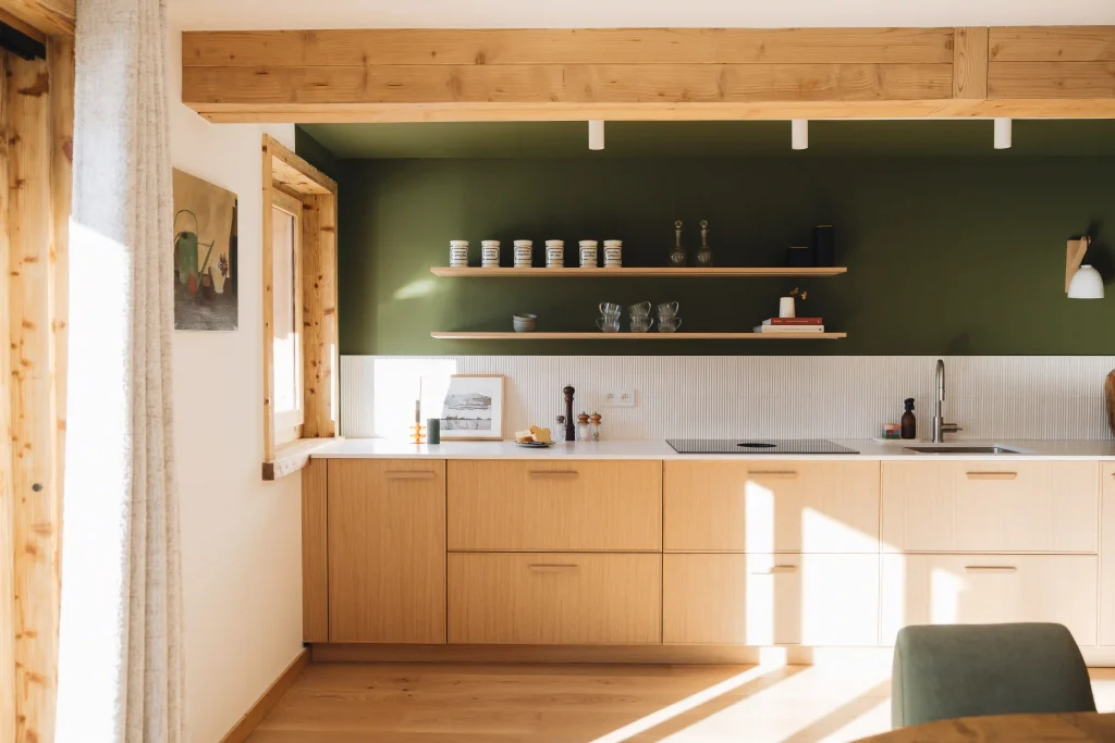 Cuisine bois Morel en chêne clair avec mur vert, étagères ouvertes, poutres apparentes et plan de travail blanc