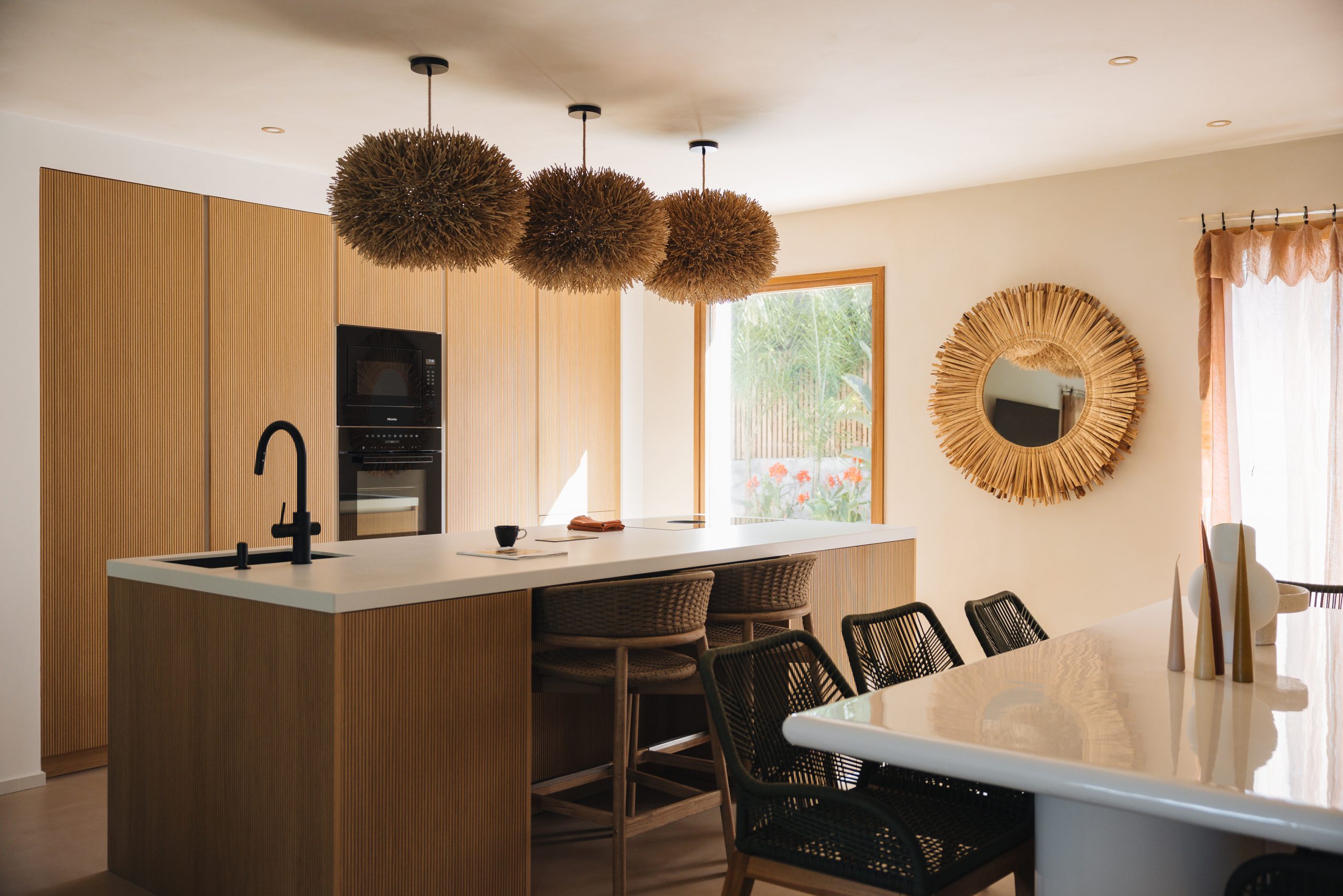 Cuisine sur-mesure Morel avec îlot central en bois cannelé, suspensions en fibres naturelles et miroir soleil en rotin